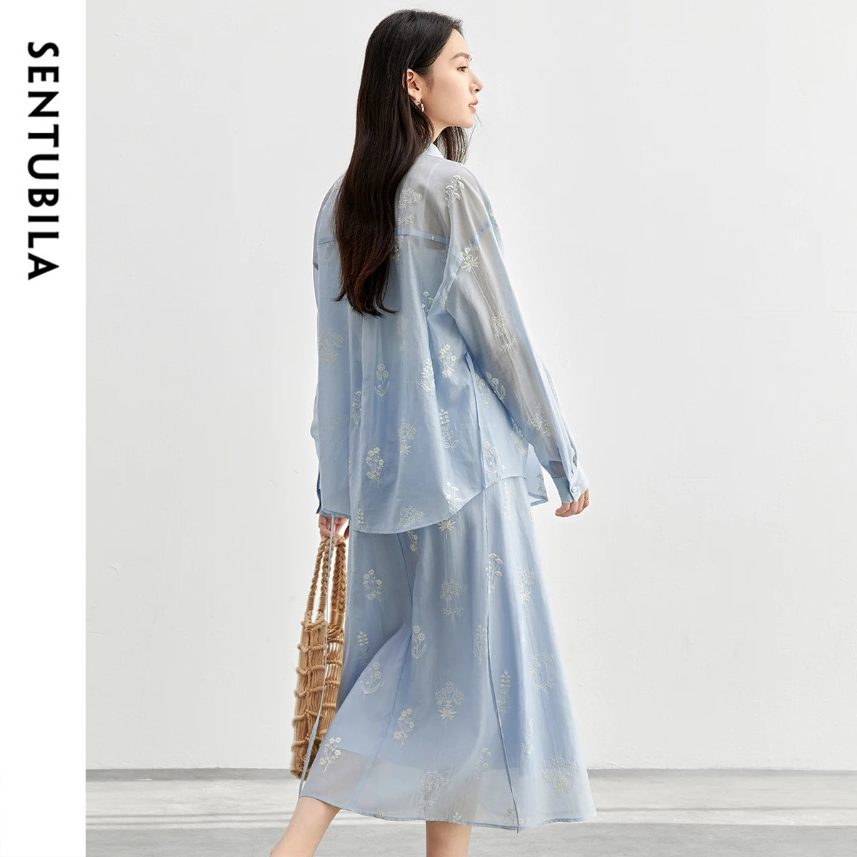 Woman wearing a light blue outfit with floral patterns, holding a woven bag on a white background.