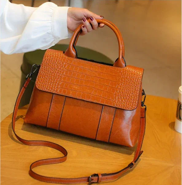 Brown leather handbag with a strap held by a person on a wooden surface.
