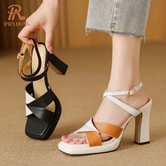 Black and white sandal held by a hand with a foot wearing a white sandal with brown accents on a beige background.