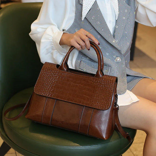 Brown leather handbag on a green chair with a person wearing a checkered blazer and white shirt.